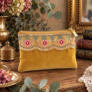 Può includere: Una pochette con cerniera in velluto giallo senape, decorata con una fascia ricamata con motivi floreali rosa, foglie verdi e paillettes dorate. La pochette presenta un bordo smerlato lungo il ricamo e un cursore della cerniera in metallo dorato.