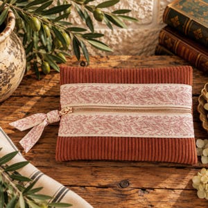 Puede incluir: Un estuche rectangular de pana color óxido con cremallera, que presenta una banda horizontal de cinta color crema con un delicado patrón de hojas rosas. El tirador de la cremallera está adornado con un lazo de tela a juego y textura.