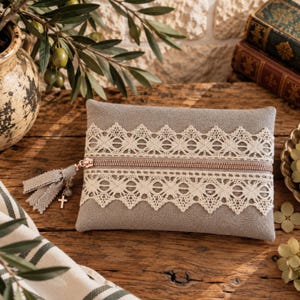 Può includere: Una pochette rettangolare in tessuto grigio con un bordo in pizzo color crema che corre orizzontalmente al centro. La pochette presenta una chiusura a zip metallica con una nappa in tessuto grigio e una piccola croce argentata sul cursore.