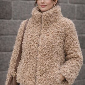 May include: A beige, textured jacket with a high collar. The jacket has a button closure and pockets. The model is wearing the jacket with white trousers and a brown handbag. The jacket is made of a soft, fuzzy material.