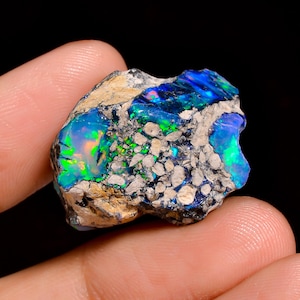 May include: A raw boulder opal specimen featuring vibrant flashes of electric blue and neon green play-of-color embedded within a textured, light tan and grey rocky matrix. The stone displays irregular, jagged edges and a natural, unpolished surface.