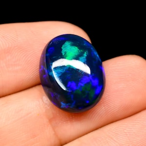 Könnte beinhalten: Ein ovaler schwarzer Opal-Edelstein mit einer polierten, glatten Oberfläche, der lebhafte Blitze in irisierendem Grün und tiefem Blau aufweist und das Licht mit einem hellen weißen Glanzlicht auf seiner abgerundeten Oberseite reflektiert.