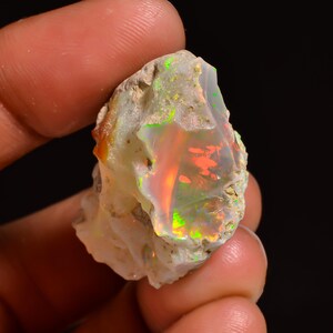 May include: A raw opal gemstone, displaying a spectrum of colours. The stone has flashes of red, green, and yellow within a milky white base. The opal is held in a hand, showing its natural, uncut form.