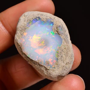 May include: A rough, uncut opal gemstone with a vibrant display of colors. The opal's interior showcases a play of iridescent hues, including blues, greens, and oranges, set against a milky white base. The stone is encased in a rough, textured matrix.