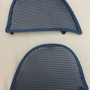 Puede incluir: Dos rejillas de altavoz de color azul oscuro con un patrón de malla perforada uniforme. Cada pieza tiene un borde superior curvo y una forma asimétrica con pequeñas pestañas de montaje que sobresalen del borde inferior para protección.