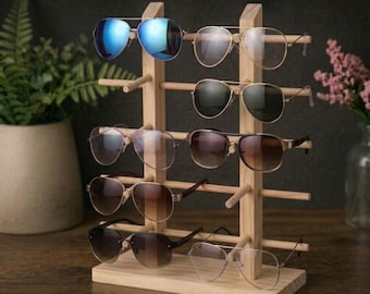 Op de afbeelding: Een houten zonnebrillenstandaard met acht zonnebrillen. De zonnebrillen hebben verschillende montuur- en lenskleuren, waaronder blauw, goud en bruin. De standaard is gemaakt van lichtgekleurd hout en heeft een eenvoudig, minimalistisch ontwerp.