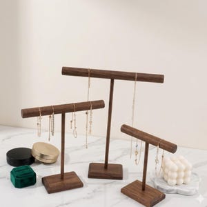 May include: Three wooden jewelry display stands in varying heights. Each stand has a horizontal bar for hanging necklaces and a square base. The stands are made of dark brown wood and are holding gold necklaces. Other jewelry boxes and decorative items are also present.
