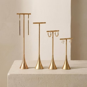 May include: Four gold-colored earring display stands of varying heights. Each stand has a slender pole and a flared base. The stands hold different styles of earrings, including long bars, hoops, and geometric shapes.