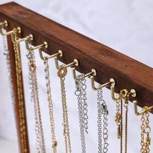 Op de afbeelding: Een houten sieradenstandaard met meerdere goudkleurige haken. Verschillende kettingen in goud en zilver hangen aan de haken. Het hout heeft een donkerbruine afwerking en de achtergrond is zacht wit.