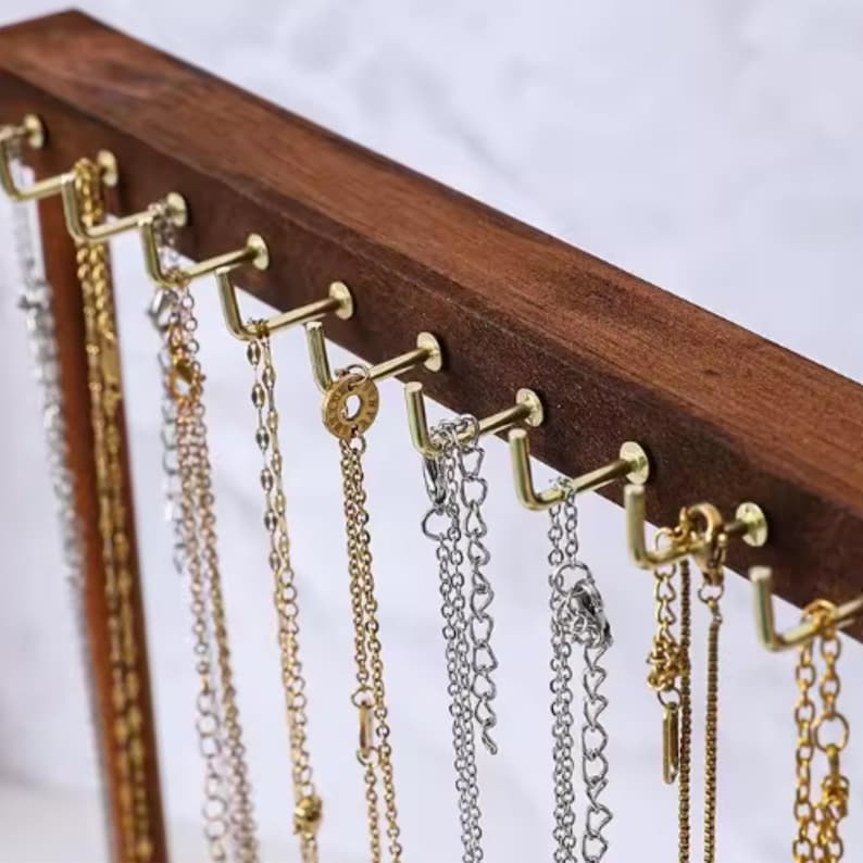 Op de afbeelding: Een houten sieradenstandaard met meerdere goudkleurige haken. Verschillende kettingen in goud en zilver hangen aan de haken. Het hout heeft een donkerbruine afwerking en de achtergrond is zacht wit.