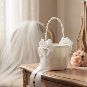 May include: Ivory-colored wedding basket with a pearl handle and white satin bows. The basket has long, flowing ribbons and is set on a wooden surface. A bouquet of pink roses is nearby, suggesting a bridal theme.
