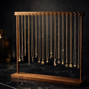 Op de afbeelding: Een houten sieradenstandaard met talrijke gouden kettingen met diverse hangerontwerpen. De kettingen hangen aan een horizontale balk en tonen een verscheidenheid aan bedels, waaronder sterren en bellen. De standaard is gemaakt van donker hout.