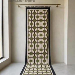 May include: A long rectangular runner rug featuring a repeating grid pattern of olive green squares on a cream background, framed by a thick black border. The rug hangs from a brass rod and displays the text Shri Kishori Exports at the top.