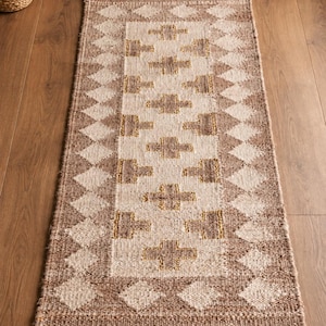 May include: A rectangular woven runner rug featuring a central panel with a repeating cross pattern in shades of tan, beige, and gold. The border consists of a diamond geometric motif in alternating light and dark brown tones with a textured finish.