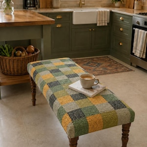 May include: A rectangular upholstered bench featuring a textured checkerboard pattern in shades of green, yellow, and gray. The bench is supported by four dark brown turned wooden legs and holds a white book with a ceramic mug on its surface.