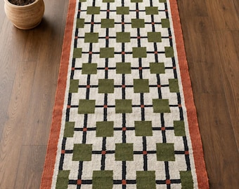 May include: A rectangular runner rug featuring a repeating grid pattern of olive green squares connected by black lines and small orange accents on a cream background, framed by a solid orange border with dark brown fringe at both ends.