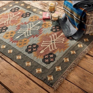 May include: A rectangular rug with a geometric pattern in grey, peach, and brown. The rug has a dark green border and a fringe. Candles and a basket with a blanket are in the background.