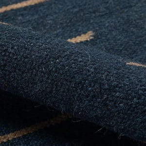 May include: Close-up of a rolled-up navy blue rug with a textured surface. The rug features a pattern of tan-colored lines. The fibers of the rug are visible, adding to its tactile appearance. The rug's design is simple and modern.