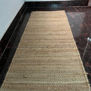 May include: A rectangular, woven jute rug in a natural tan color. The rug has a simple, textured design and is placed on a dark, patterned floor. The rug is a long, narrow runner, suitable for hallways or entryways.