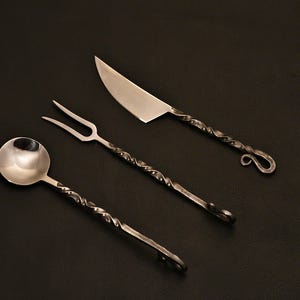 May include: A set of three handcrafted metal serving utensils: a spoon, a fork, and a knife. Each piece features a twisted handle design and a polished silver finish. The knife has a curved blade.