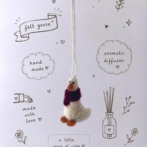 Puede incluir: Adorno de ganso de fieltro hecho a mano con cuerpo blanco, patas naranjas y bufanda burdeos. El adorno está suspendido por un cordón blanco. La imagen incluye el texto "felt goose", "hand made" y "a little piece of calm".
