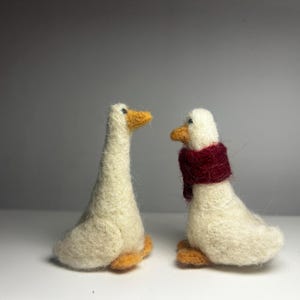 Puede incluir: Dos gansos de fieltro hechos a mano. Un ganso se alza con pico y patas amarillas. El otro ganso está sentado y lleva una bufanda burdeos. Ambos gansos tienen cuerpos blancos.