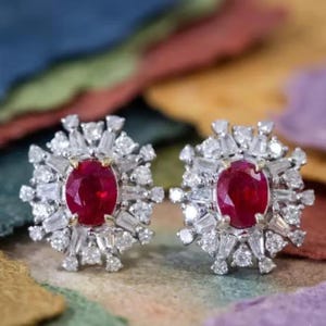 Puede incluir: Un par de pendientes de botón con rubíes rojos de talla ovalada engastados en metal plateado. Cada pendiente presenta un rubí central rodeado por un halo de diamantes blancos transparentes de talla baguette y redonda alternados.