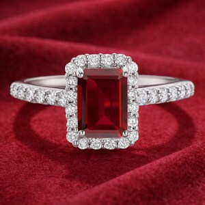 Puede incluir: Un anillo de plata con una gema rectangular de color rojo intenso, rodeada por un halo de gemas claras más pequeñas. La banda del anillo también está adornada con pequeñas gemas claras. El anillo se muestra sobre un fondo rojo.