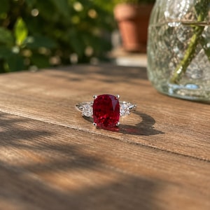 Può includere: Anello d'argento con una grande gemma rosso scuro a taglio cuscino come punto focale. Due gemme più piccole, a taglio quadrato, affiancano la pietra principale. L'anello è appoggiato su una superficie di legno.
