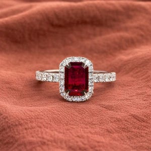 May include: A silver ring with a large, rectangular, deep red gemstone at the center, surrounded by a halo of smaller, clear stones. The band is also adorned with small, clear stones. The ring is set against a reddish-brown fabric background.