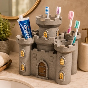 May include: A grey castle-shaped toothbrush holder with a textured stone finish, featuring a central arched wooden door, multiple towers, and small windows with warm yellow light. It holds a tube of Oral-B toothpaste and four toothbrushes.