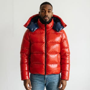 Puede incluir: Una chaqueta acolchada de color rojo brillante con cuello alto, cremallera frontal negra y capucha extraíble con forro interior azul marino oscuro. La prenda presenta paneles acolchados horizontales y se lleva sobre unos vaqueros azules.