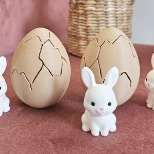 May include: Easter decorations. Two large, tan-coloured eggs with a cracked design and three small white bunny figurines with pink noses. The items are on a pink surface, with a woven basket in the background.