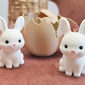 May include: Two white bunny figurines with black eyes and pink noses sit on a pink surface. A cracked eggshell and a purple Easter egg are behind them. The bunnies are part of an Easter-themed decoration.