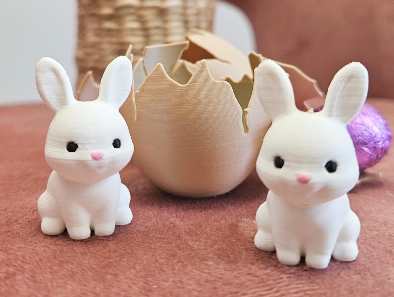 May include: Two white bunny figurines with black eyes and pink noses sit on a pink surface. A cracked eggshell and a purple Easter egg are behind them. The bunnies are part of an Easter-themed decoration.