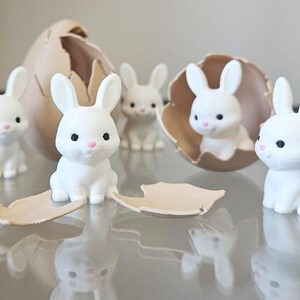 May include: A group of white rabbit figurines with black eyes and pink noses. Some are inside a tan eggshell, others are standing. The figurines are on a reflective surface, creating a mirrored effect.