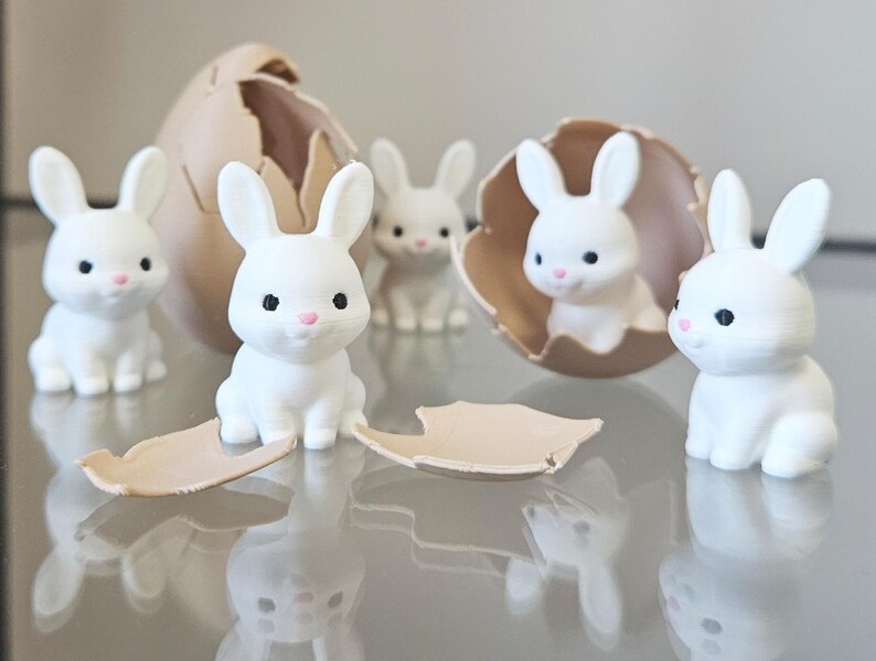 May include: A group of white rabbit figurines with black eyes and pink noses. Some are inside a tan eggshell, others are standing. The figurines are on a reflective surface, creating a mirrored effect.