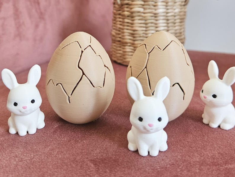 May include: Easter decorations. Two large, tan-coloured eggs with a cracked design and three small white bunny figurines with pink noses. The items are on a pink surface, with a woven basket in the background.