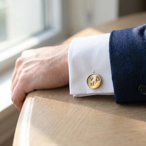 Puede incluir: Gemelos dorados con un diseño circular que presenta un ciervo y árboles. Los gemelos están sujetos a un puño de camisa blanca, combinados con una blazer azul marino. La imagen es un primer plano, destacando los detalles de los accesorios.