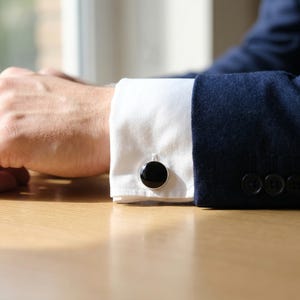 May include: Close-up of a white dress shirt cuff with a black onyx cufflink. The cufflink is round with a silver-colored border. A navy blue suit jacket is visible in the background.