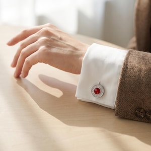 May include: A circular silver-toned cufflink featuring a vibrant red center stone surrounded by a border of small, clear, sparkling crystals. The cufflink is fastened to the crisp white cuff of a long-sleeved shirt worn under a brown blazer.