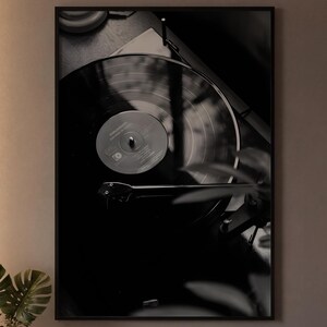 Op de afbeelding: Een ingelijste zwart-wit wandkunstprint met een vinylplaat op een platenspeler. Het kunstwerk heeft een compositie met hoog contrast, diepe schaduwen en highlights, waarbij de groeven van de plaat en de toonarm duidelijk zichtbaar zijn.