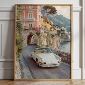 May include: A framed wall art print depicting a white vintage sports car driving on a coastal road. The scene features a pink building with an arched stone tunnel, lush greenery, and a mountain landscape overlooking the sea. The car has a license plate reading PALMS CLUB.