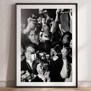 May include: A black and white framed wall art print depicting a crowd of photographers holding cameras and flashes up to their faces. The scene captures multiple individuals in dark clothing, each focused on capturing images with various camera models.