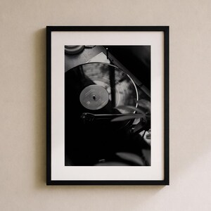 Op de afbeelding: Een zwart-wit ingelijste wandkunstprint met een bovenaanzicht van een vinylplaat die op een platenspeler draait. De compositie toont de cirkelvormige groeven van de plaat, het centrale label en de toonarm die op het oppervlak rust.
