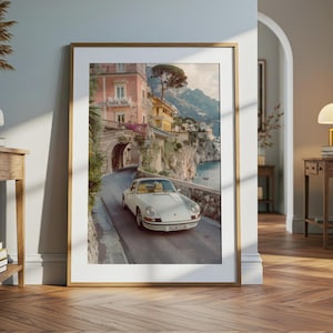 May include: A framed wall art print featuring a white vintage sports car driving on a coastal road. The scene includes pastel-colored buildings, a stone archway, and cliffs overlooking the sea. The car license plate reads PALMS CLUB.