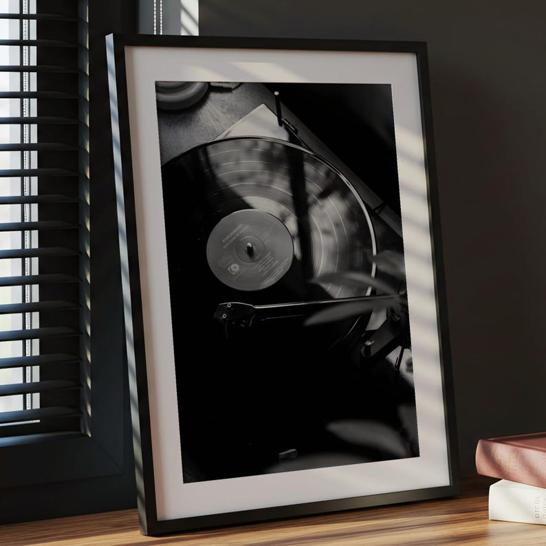 Op de afbeelding: Een zwart-wit ingelijste print met een contrastrijke foto van een vinylplaat die op een platenspeler draait. De afbeelding toont de cirkelvormige groeven van de plaat, het centrale label en de toonarm, geplaatst in een dunne zwarte lijst.