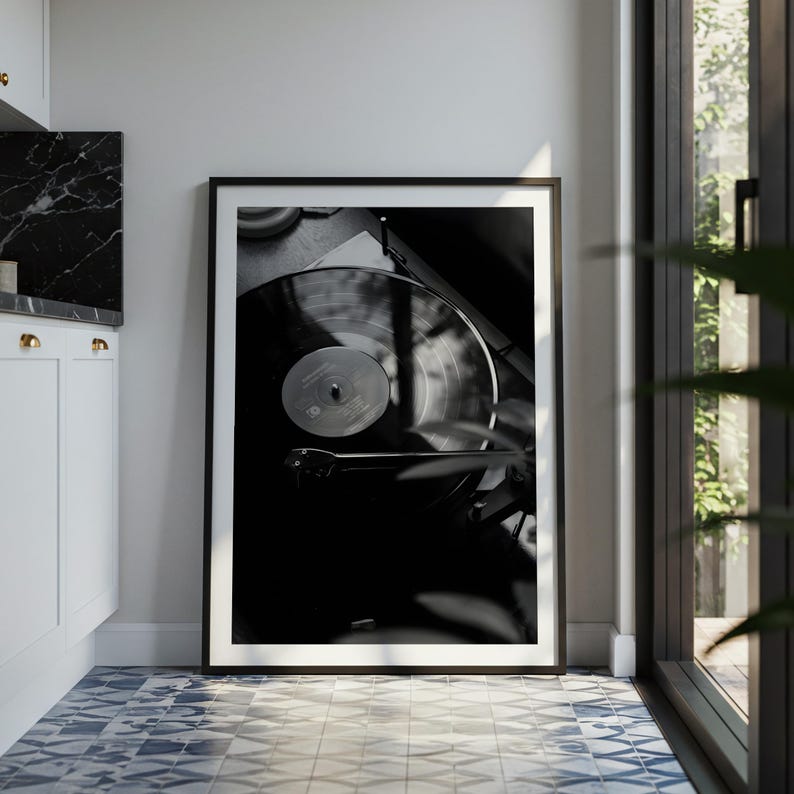 Op de afbeelding: Een zwart-wit ingelijste wandkunstprint met een contrastrijke foto van een vinylplaat die op een platenspeler draait. De afbeelding toont de cirkelvormige groeven van de plaat, het centrale label en de toonarm in een dunne zwarte lijst.