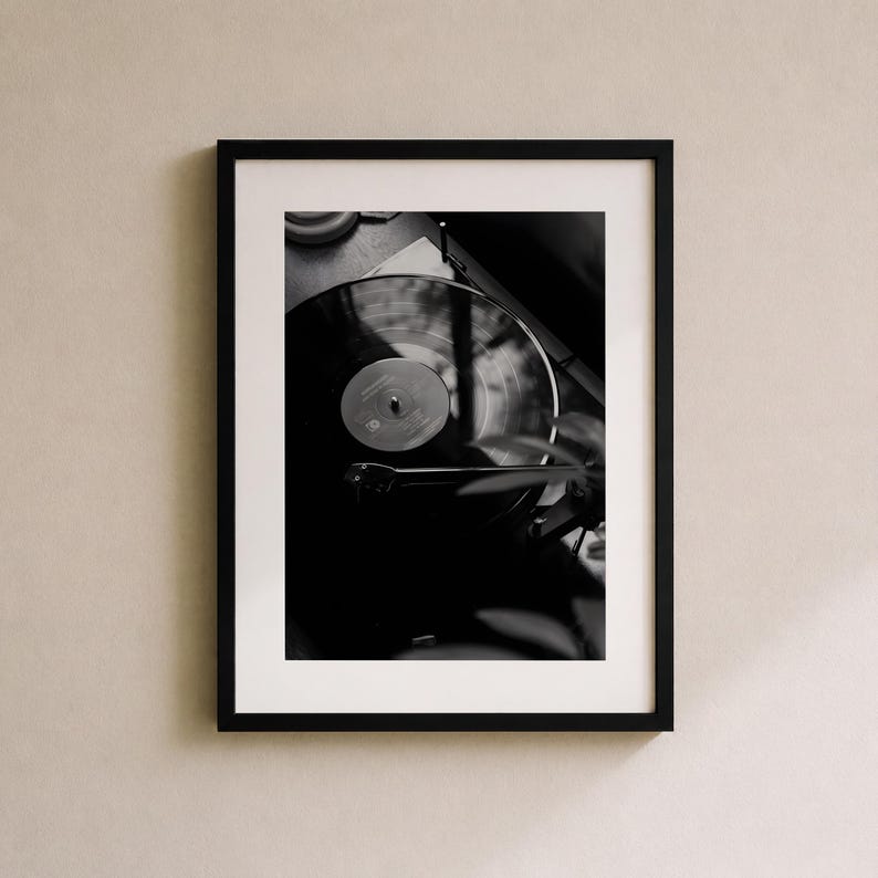 Op de afbeelding: Een zwart-wit ingelijste wandkunstprint met een bovenaanzicht van een vinylplaat die op een platenspeler draait. De compositie toont de cirkelvormige groeven van de plaat, het centrale label en de toonarm die op het oppervlak rust.