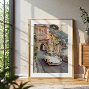 May include: A framed wall art print featuring a white vintage sports car driving on a coastal road. The scene includes pastel-colored buildings, a stone archway, and a cliffside overlooking the sea. The car has a license plate reading PALMO CLUB.